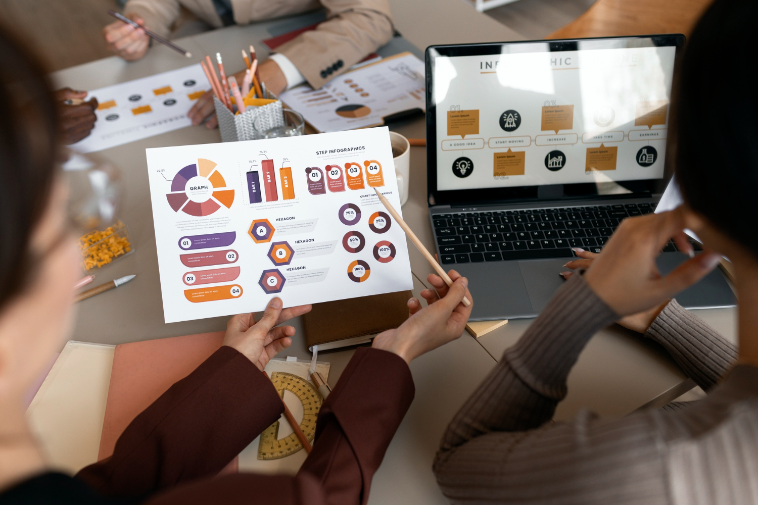 Overhead view of colleagues collaborating at an office desk, pointing to a printed sheet of colorful infographics, bar charts, and diagrams beside an open laptop displaying workflow slides.